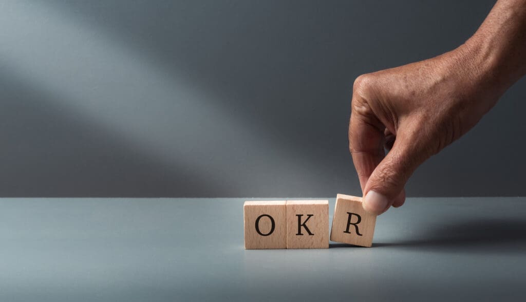 A hand placing wooden blocks on a table with the letters OKR. Credit: https://www.istockphoto.com/portfolio/robatthepub?mediatype=photography