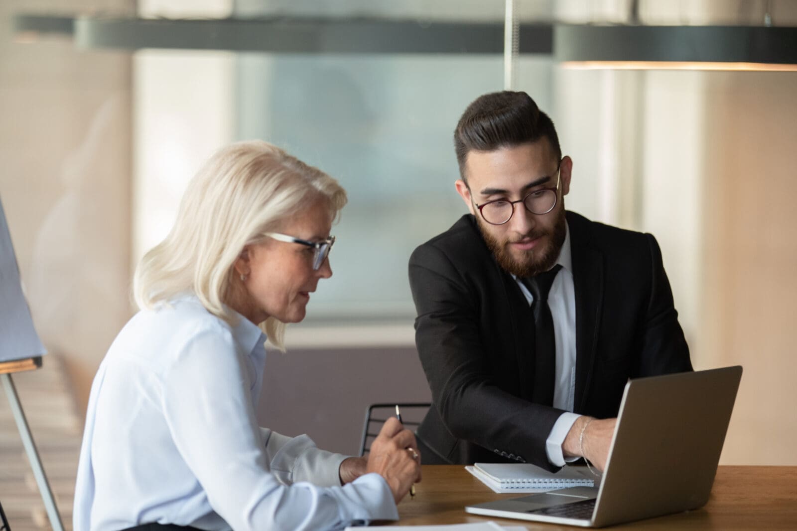 Millennial Arabian male employee point at laptop screen discussing ideas with mature female colleague
